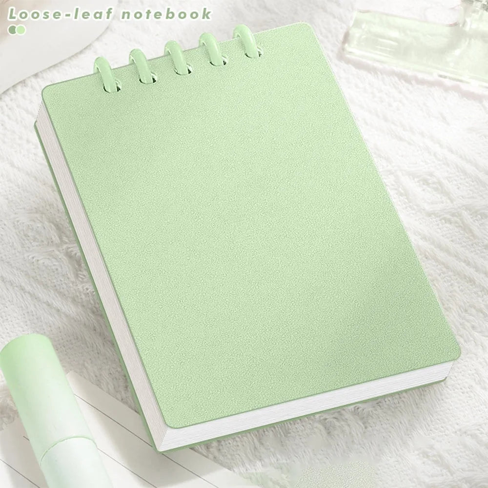 Cute Small notepad with light Pastel green cover and lined pages on a light wooden surface with magazines underneath.