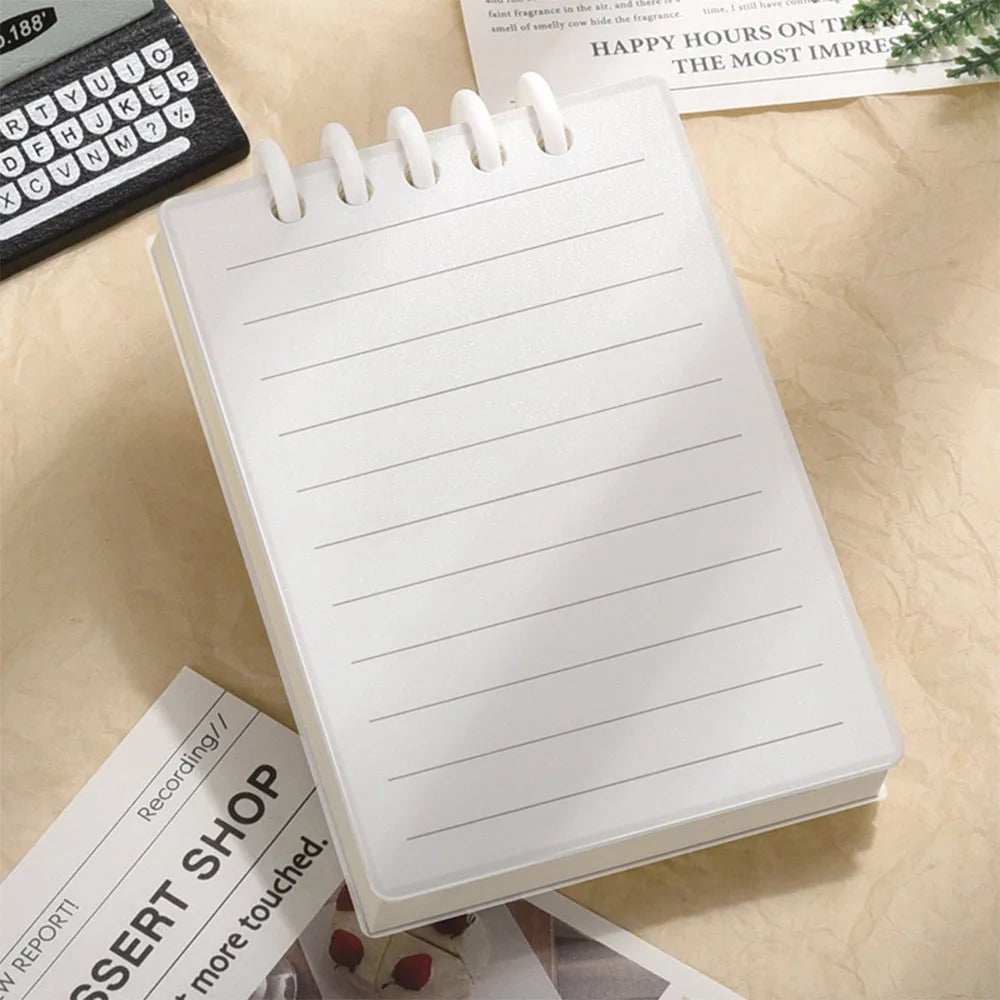 Cute Small notepad with translucent cover and lined pages on a light wooden surface with magazines underneath.