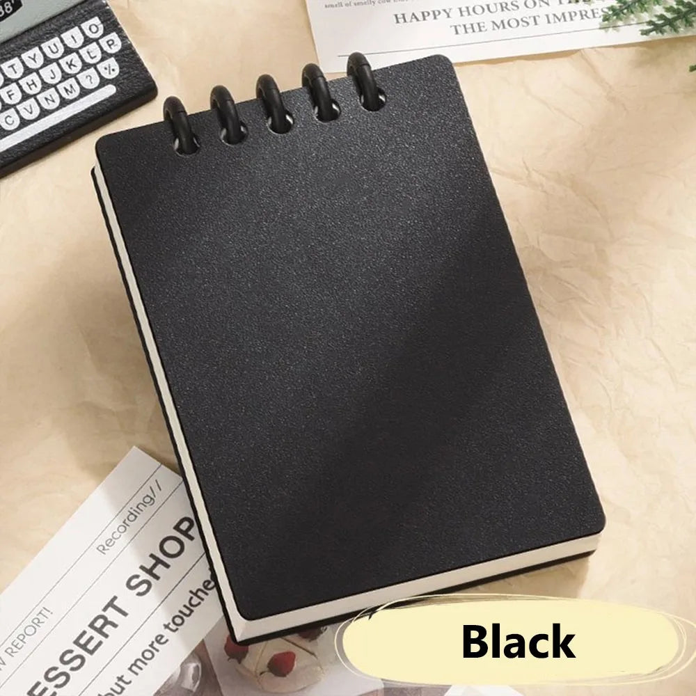 Black spiral-bound notebook on a light surface with text and a keyboard in the background