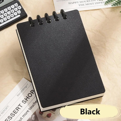 Black spiral-bound notebook on a light surface with text and a keyboard in the background