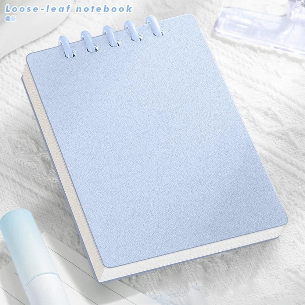 Cute Small notepad with light Pastel blue cover and lined pages on a light wooden surface with magazines underneath.