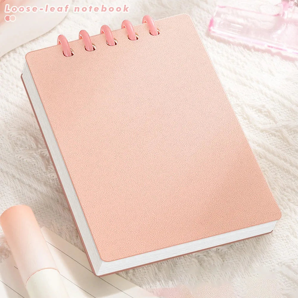 Cute Small notepad with light Pastel pink cover and lined pages on a light wooden surface with magazines underneath.