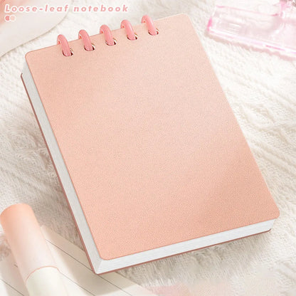 Cute Small notepad with light Pastel pink cover and lined pages on a light wooden surface with magazines underneath.
